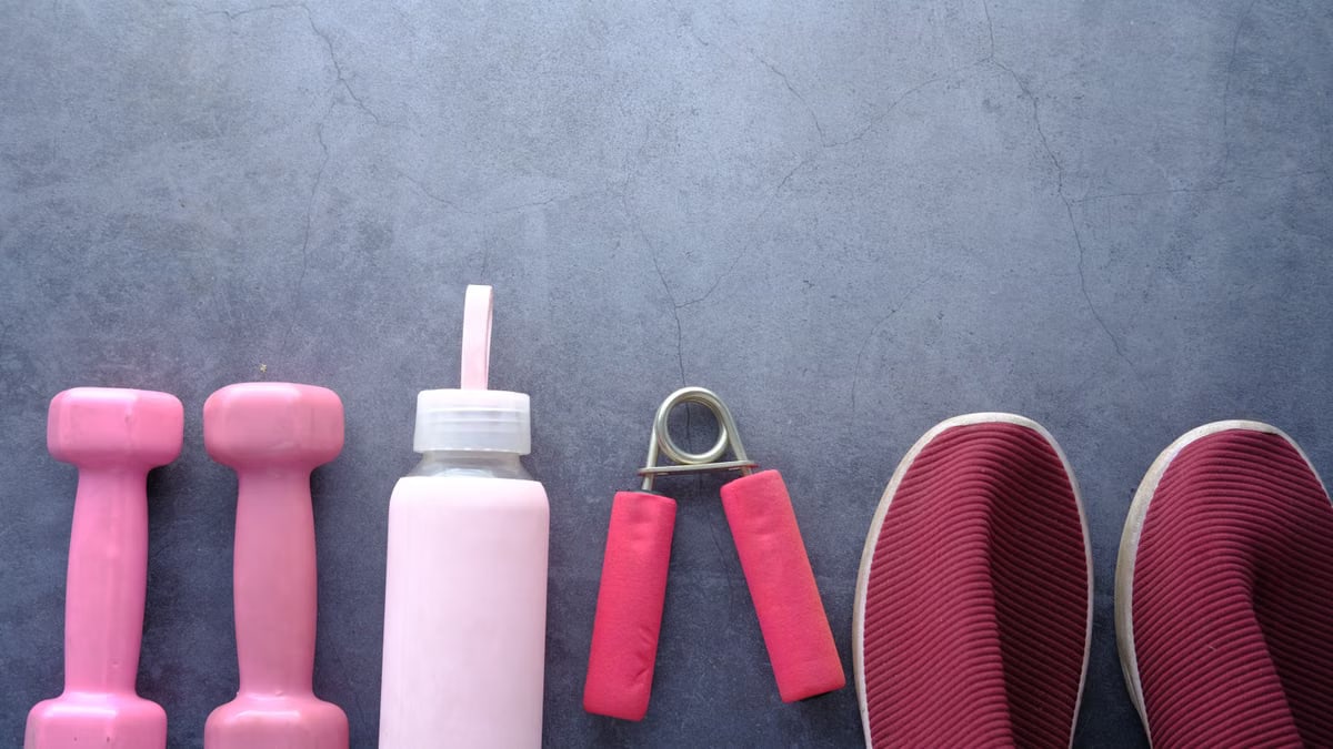 Flat lay of pink fitness equipment and shoes on a gray surface including dumbbells, water bottle, and grip strengthener.