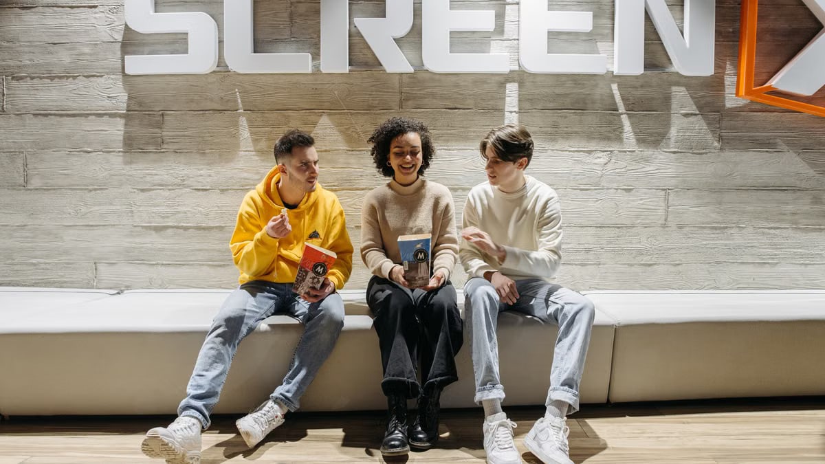 Three young adults sitting under cinema signage, enjoying snacks and conversation.